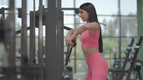 A-hispanic-brunette-woman-in-a-pink-suit-pulls-a-rope-in-a-crossover-with-her-hands-to-train-her-shoulders.-Shoulder-workout-in-a-trainer.-Professional-woman-instructor